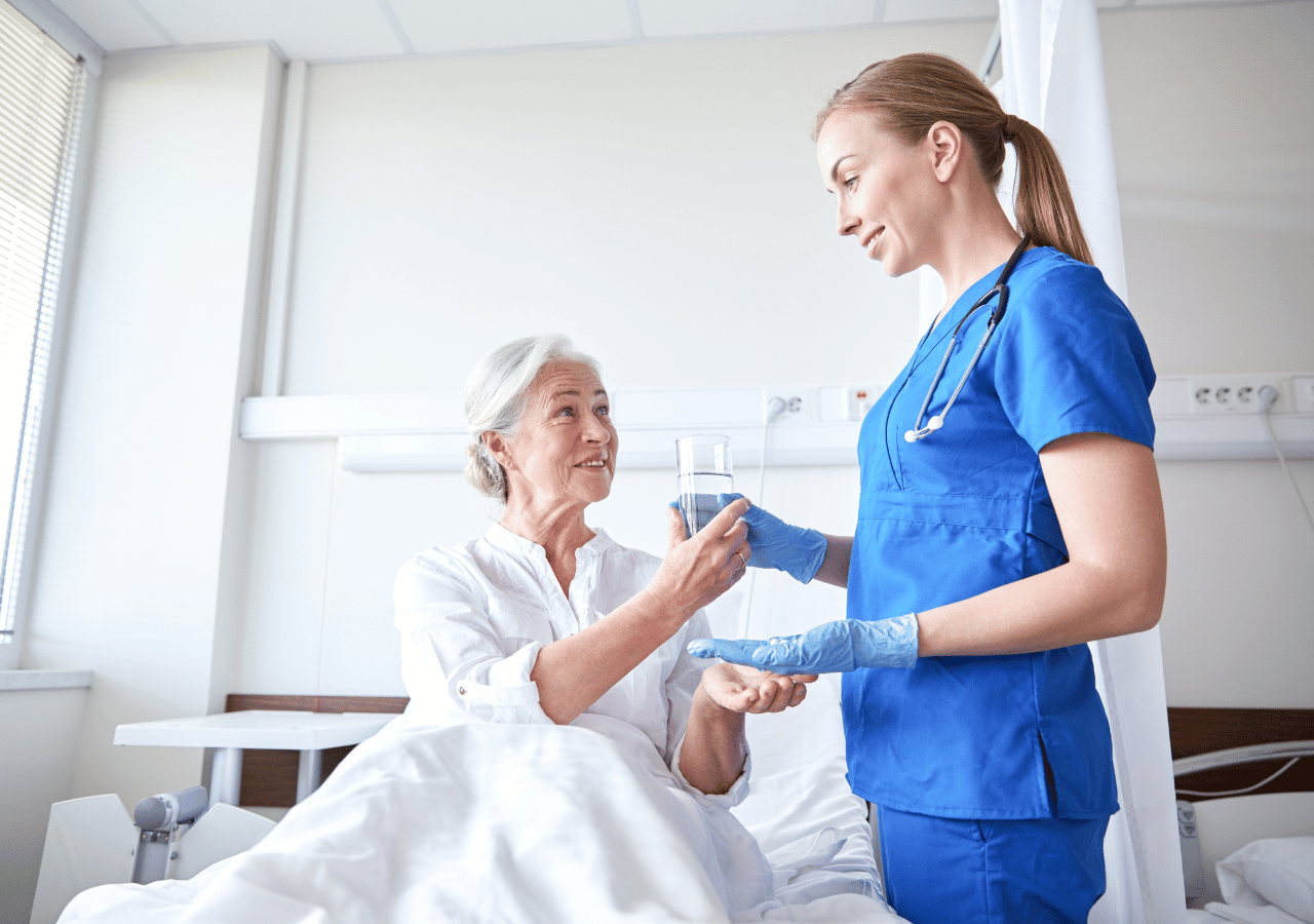 A nurse in blue scrubs with a stethoscope helps an elderly female patient in a white hospital gown drink from a glass of water in a bright hospital room.
