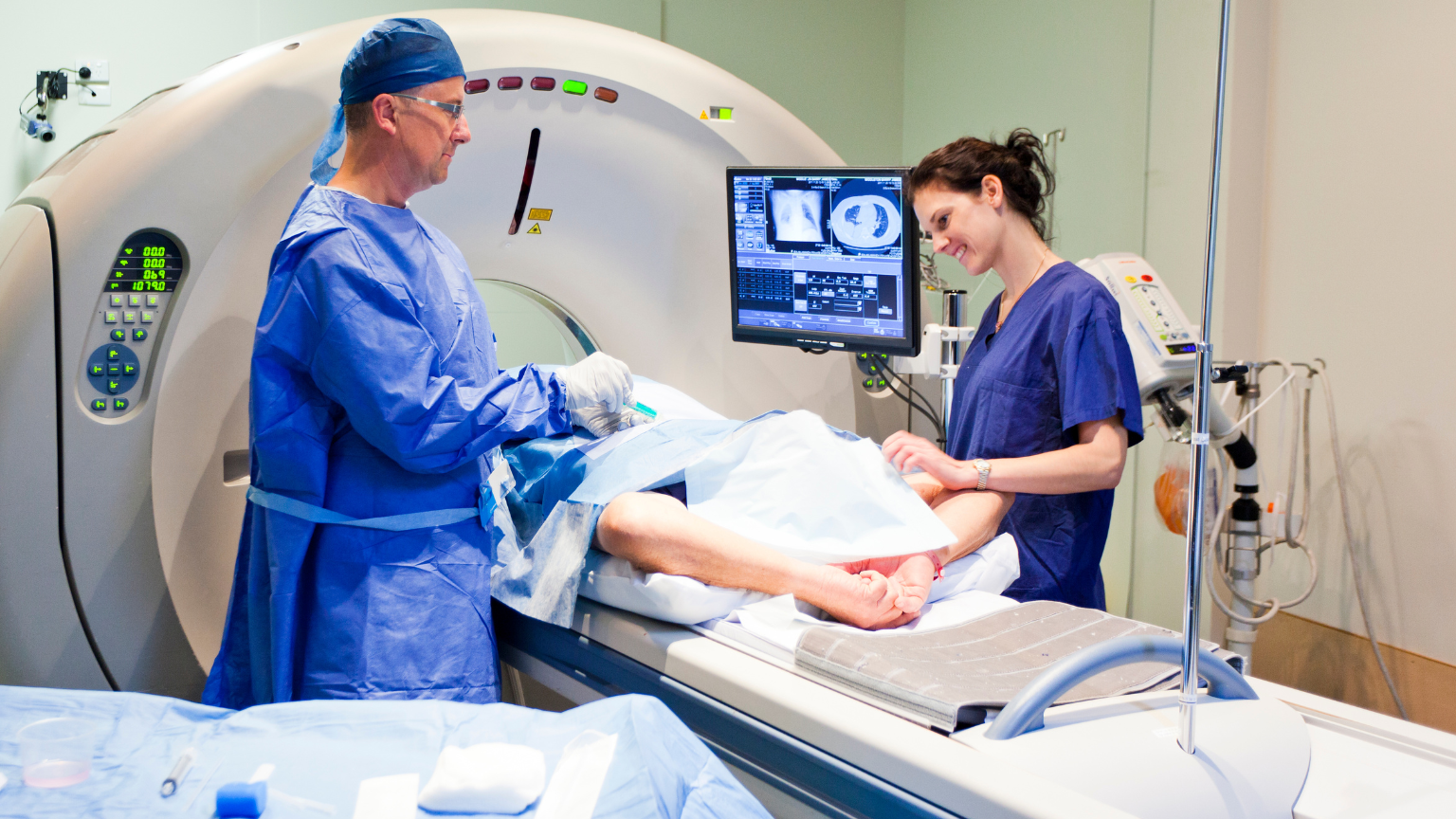 Two medical professionals in surgical scrubs attend to a patient on a CT scanner table, with medical imaging displays visible in the background showing scan results.