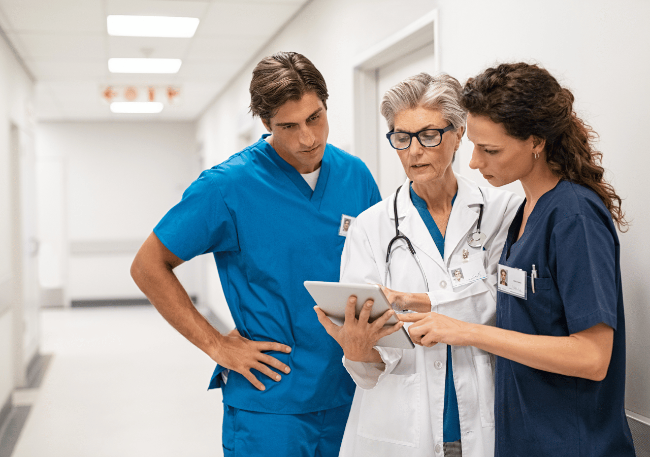 Three healthcare professionals in scrubs and a white coat consult together while looking at a tablet in a bright hospital hallway.