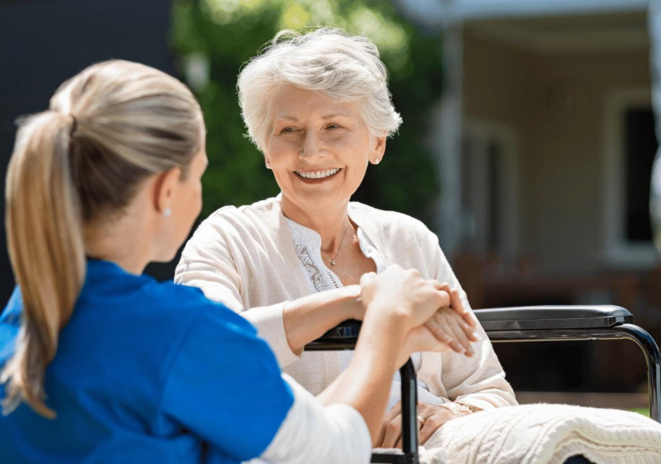 A healthcare worker in blue scrubs holds hands with an elderly woman in a wheelchair, both smiling warmly during what appears to be a compassionate care moment outdoors near a residential building.