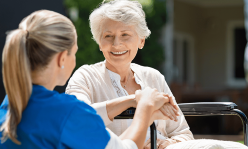 A healthcare worker in blue scrubs holds hands with an elderly woman in a wheelchair, both smiling warmly during what appears to be a compassionate care moment outdoors near a residential building.