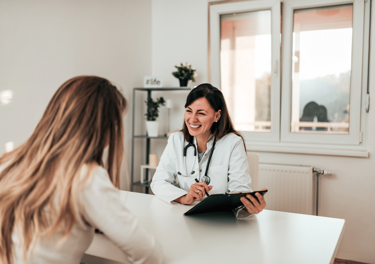 A doctor wearing a white coat and stethoscope sits at a desk with a tablet, smiling while consulting with a blonde patient in a bright medical office.