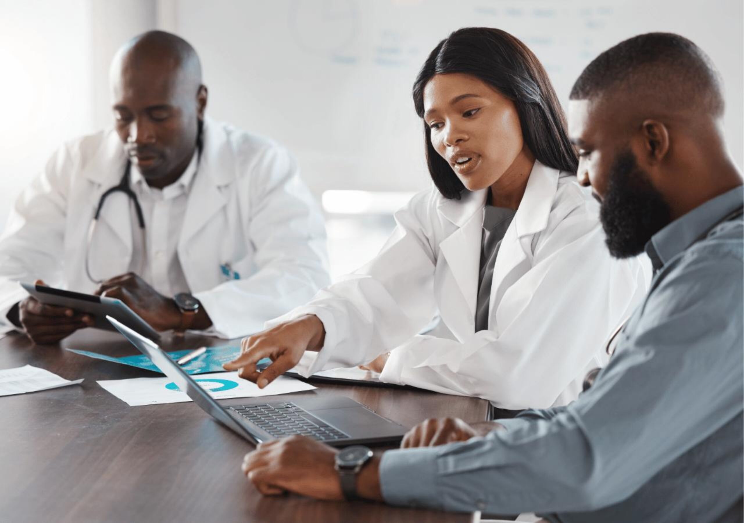 Three healthcare professionals in white coats collaborate around a laptop and documents during a medical consultation or meeting, with one doctor wearing a stethoscope and another person in business attire participating in the discussion.