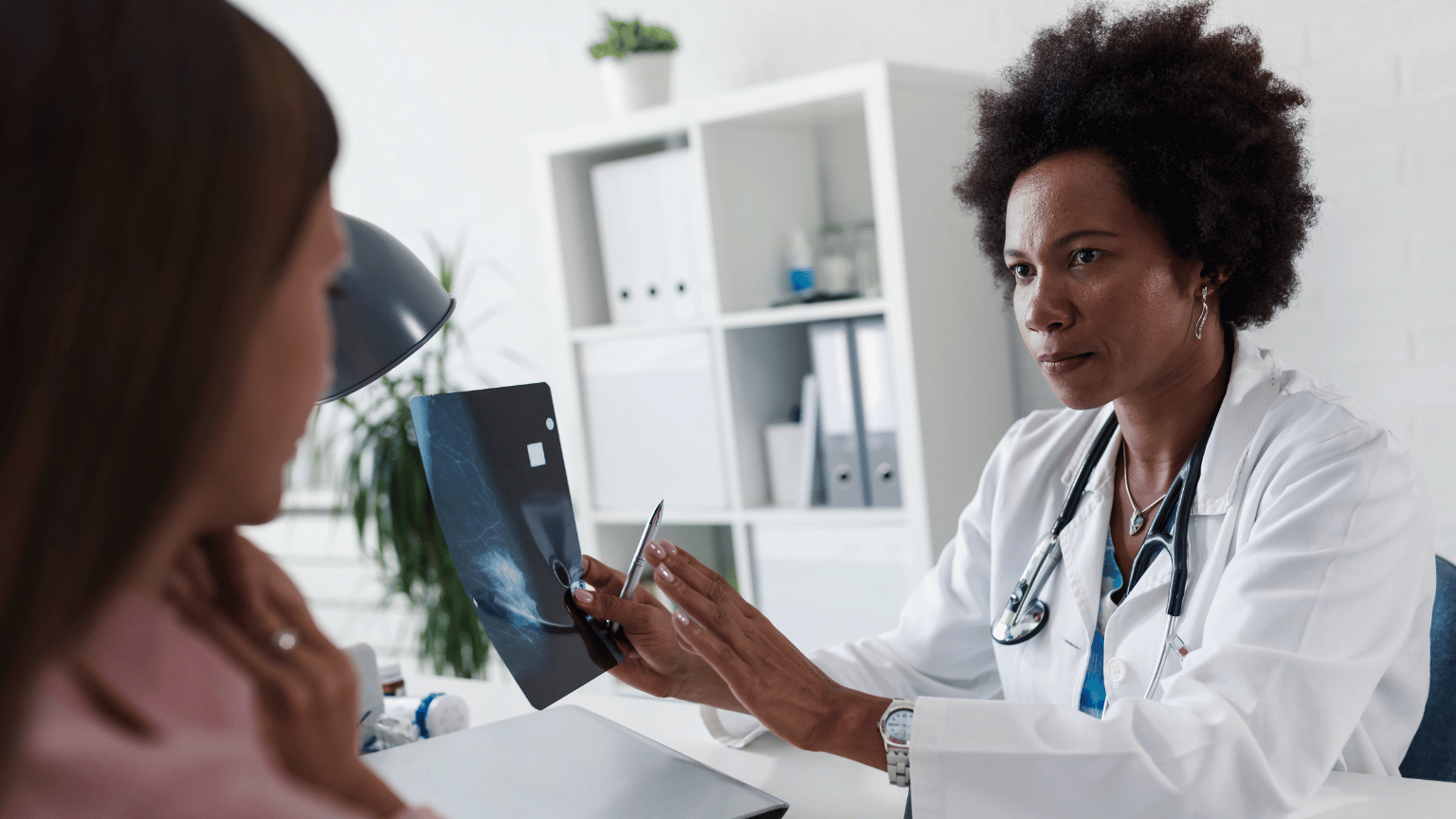 A doctor with curly hair wearing a white coat and stethoscope shows an X-ray image to a patient during a medical consultation in a modern clinical office.