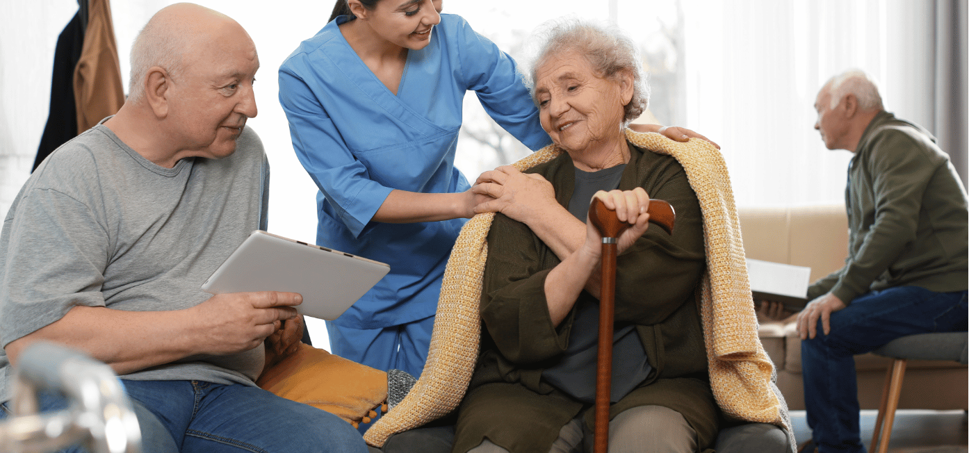 A healthcare worker in blue scrubs provides care to an elderly woman with a cane and blanket while a man with a tablet sits nearby in what appears to be a senior living facility or care home.