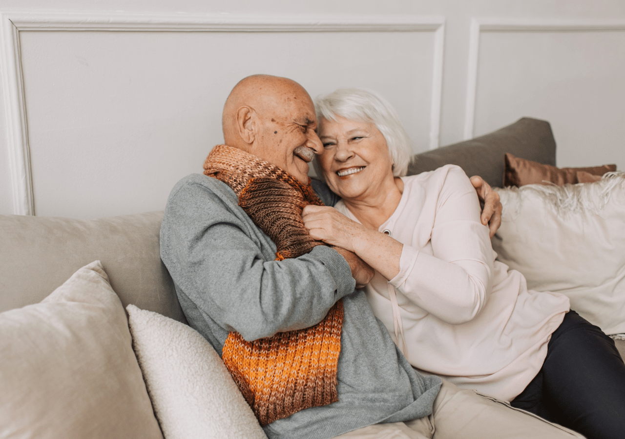 An elderly couple sits closely together on a beige couch, smiling and embracing warmly. The man wears a gray sweater with an orange and brown knit scarf, while the woman has white hair and wears a light pink top.