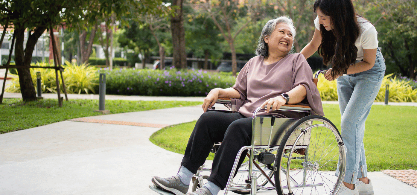 An elderly woman in a wheelchair smiles while a younger woman stands beside her with a caring touch on her shoulder, both enjoying time together in a park setting with green lawns and trees.