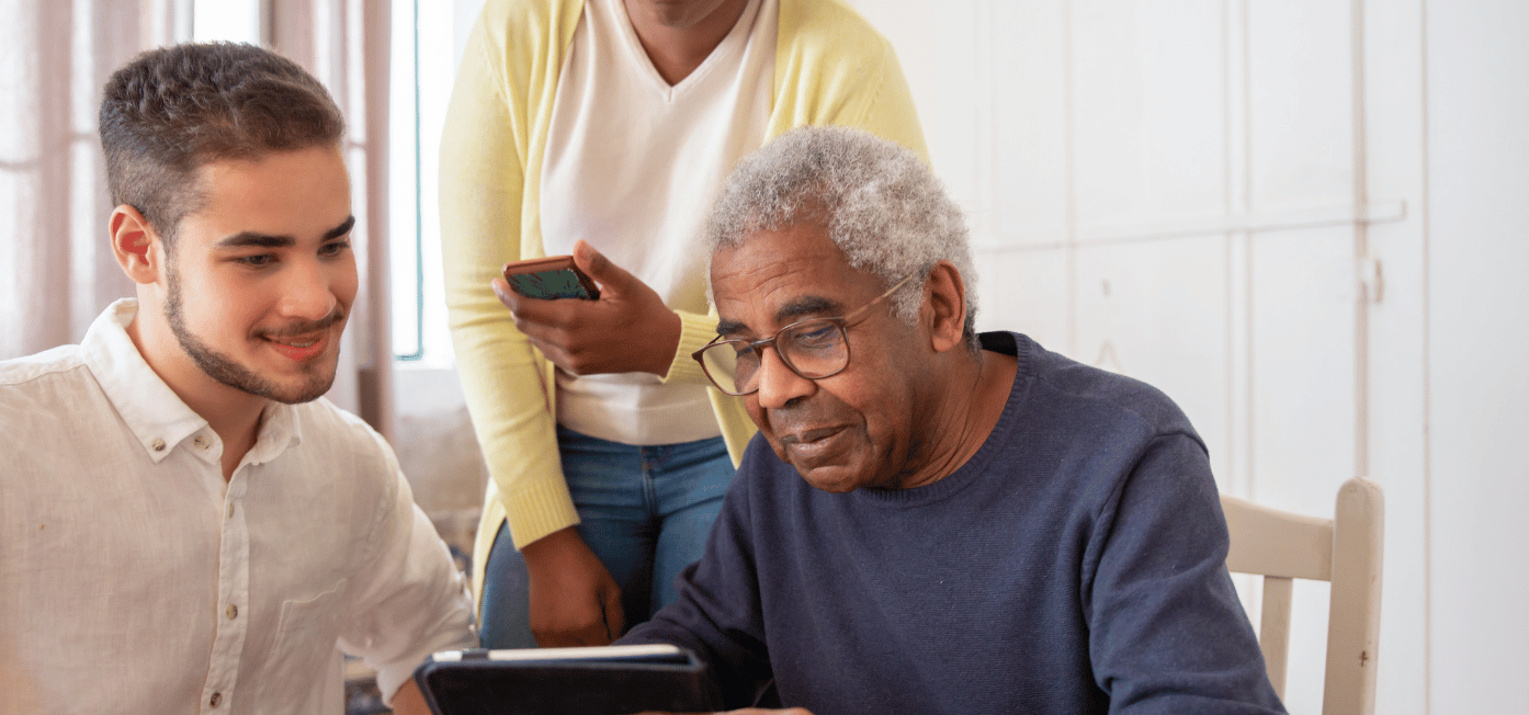 A young man in a white shirt sits beside an older man with glasses and gray hair who is looking at a tablet, while a woman in a yellow cardigan stands behind them holding a smartphone in a bright, home-like setting.
