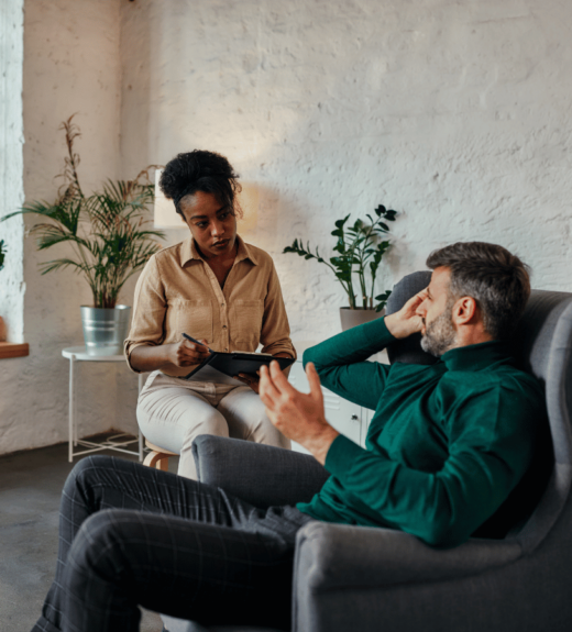 A therapist or counselor sits on a chair taking notes while speaking with a male client who is seated in a gray armchair with his hand on his forehead in a modern office setting with white brick walls and plants.