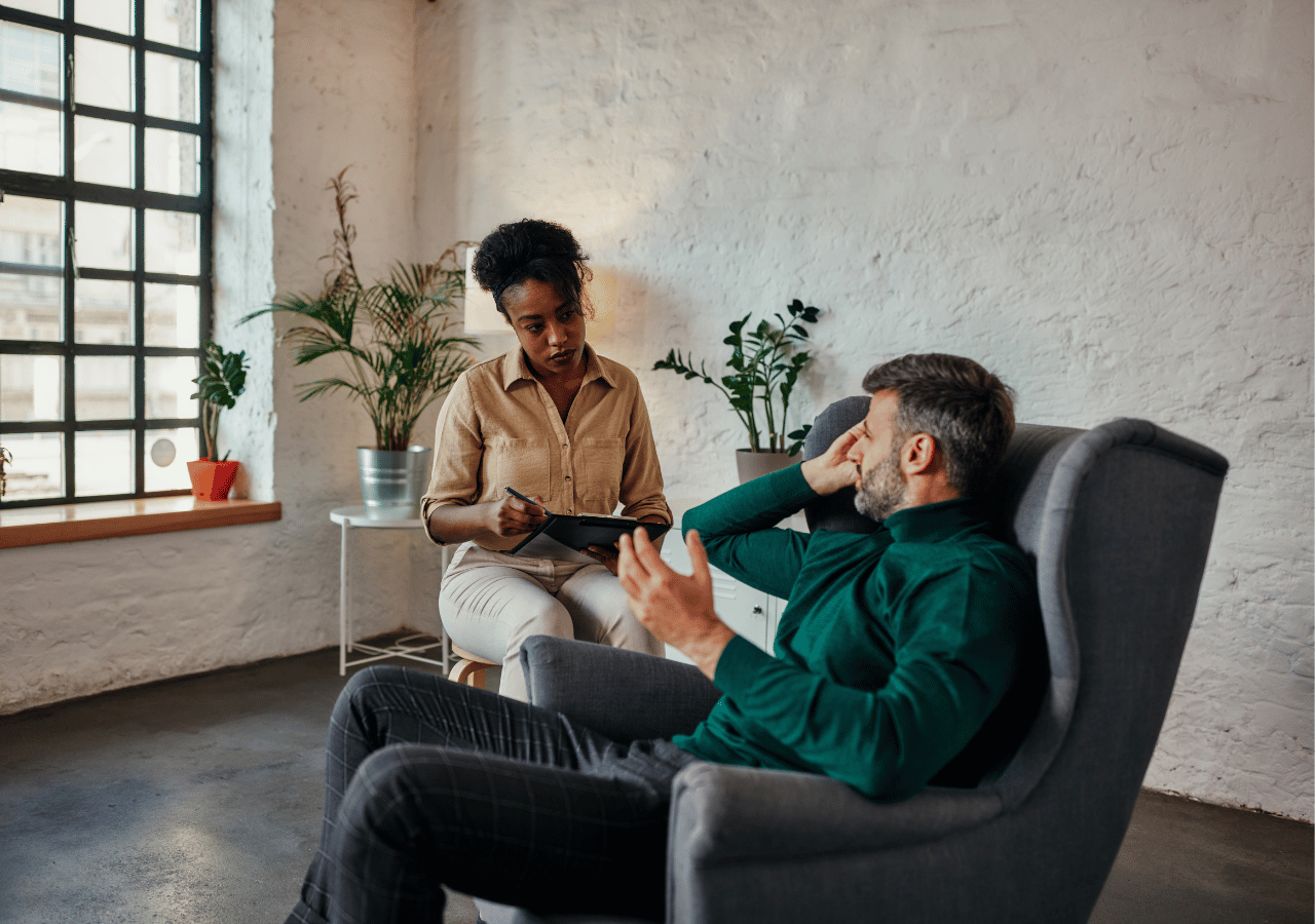 A therapist or counselor sits on a chair taking notes while speaking with a male client who is seated in a gray armchair with his hand on his forehead in a modern office setting with white brick walls and plants.