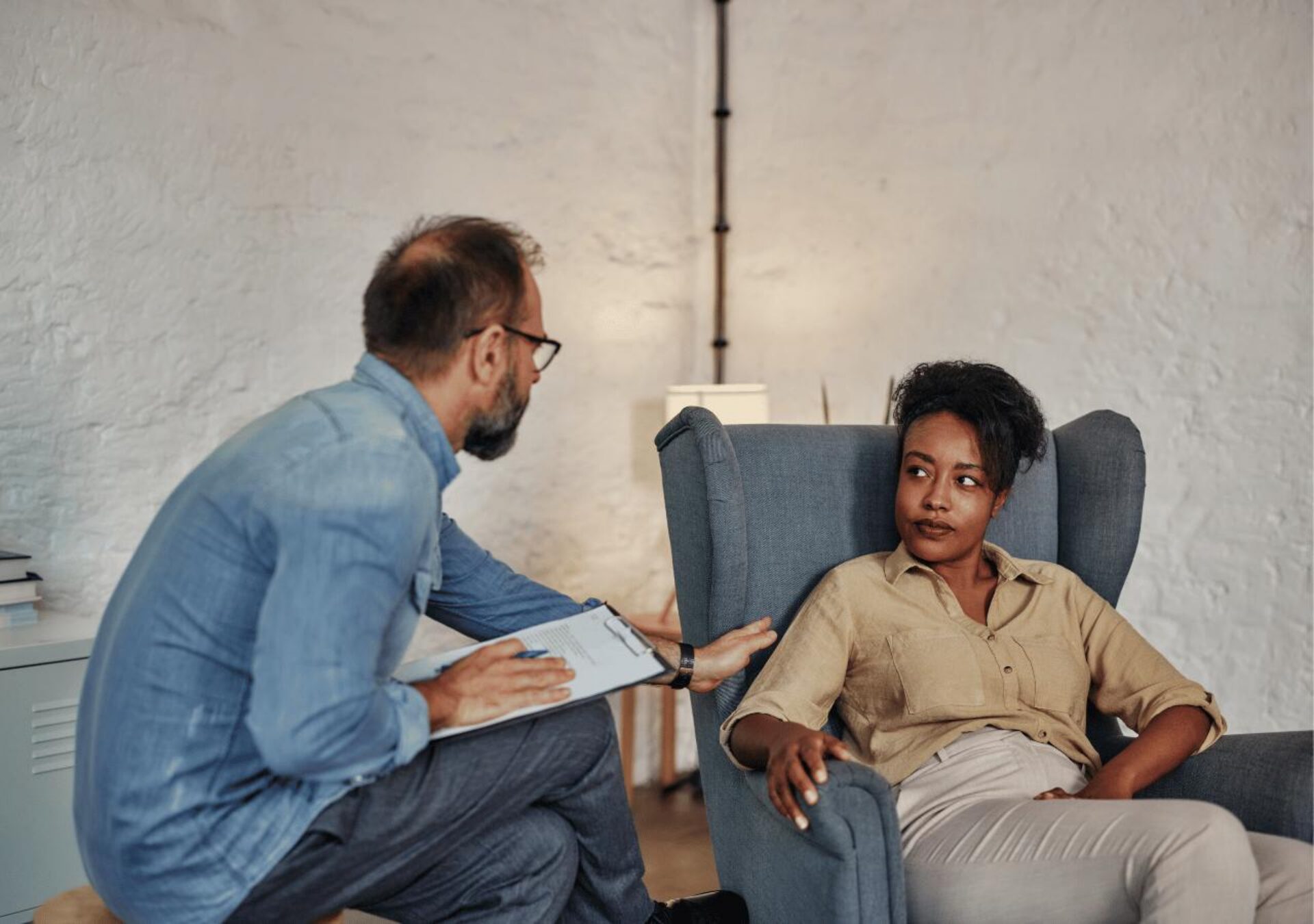 A therapist or counselor with glasses and a blue shirt sits on a chair taking notes while speaking with a Black woman client who is seated in a comfortable blue armchair in what appears to be a therapy office setting.