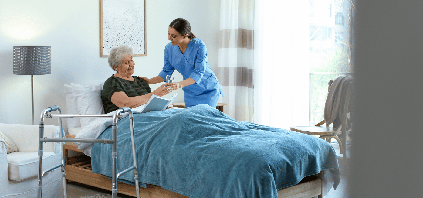 A healthcare worker in blue scrubs assists an elderly patient in bed, helping her with paperwork or medical documents in a bright, modern healthcare room with a walker nearby.