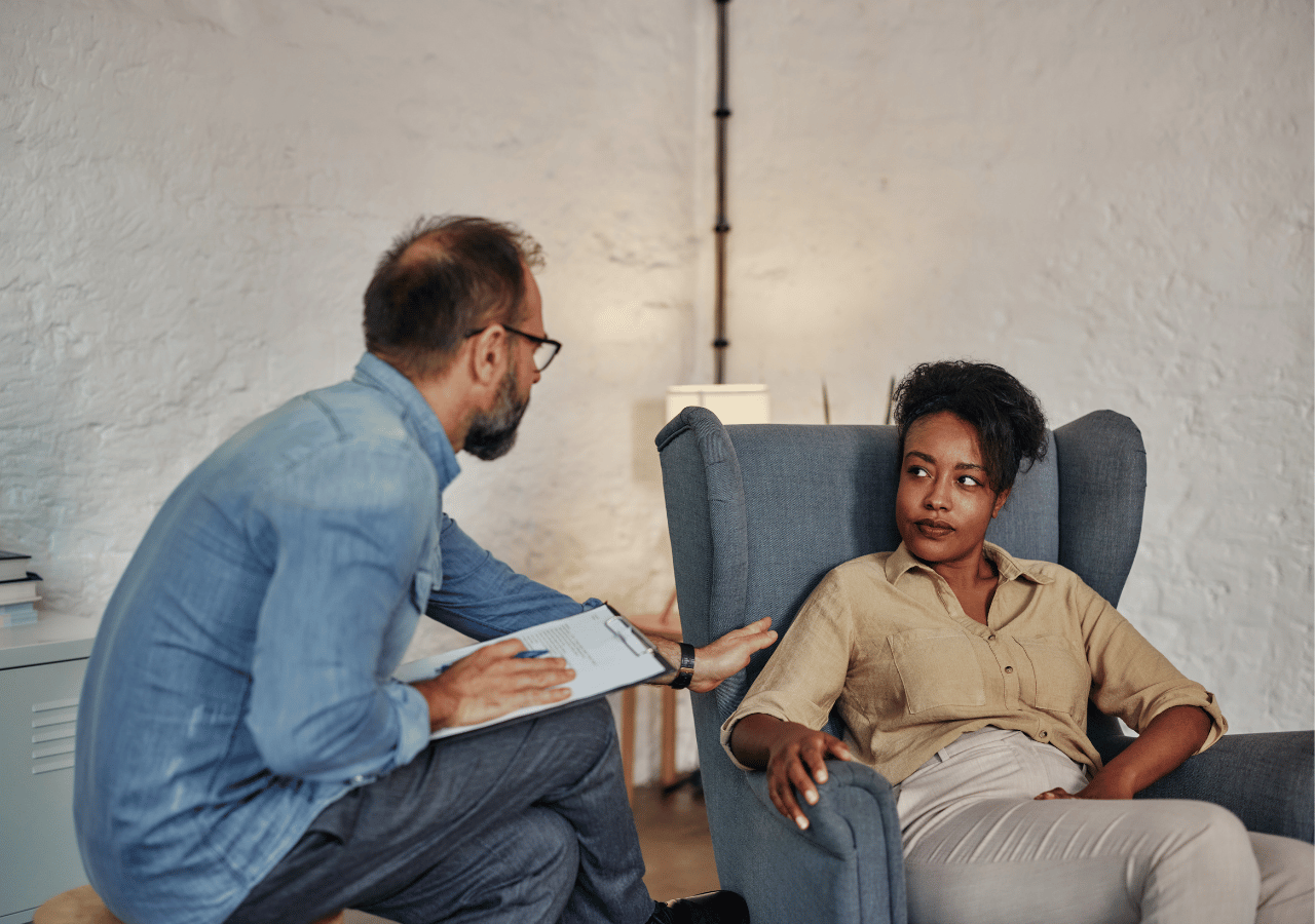A therapist or counselor with glasses and a blue shirt sits on a chair taking notes while speaking with a Black woman client who is seated in a comfortable blue armchair in what appears to be a therapy office setting.
