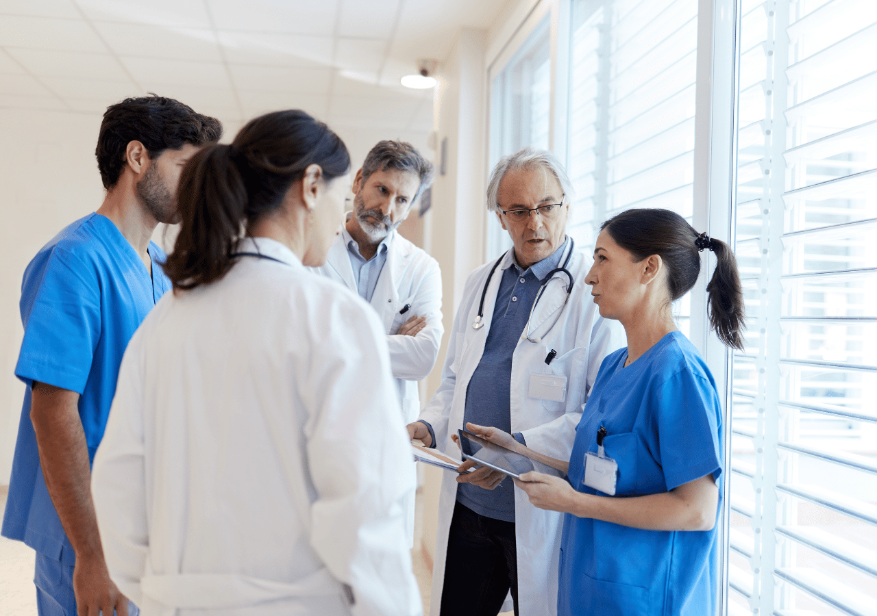 A group of five medical professionals in white coats and blue scrubs having a discussion in a hospital hallway, with one doctor holding a tablet while they appear to be reviewing patient information or medical cases.