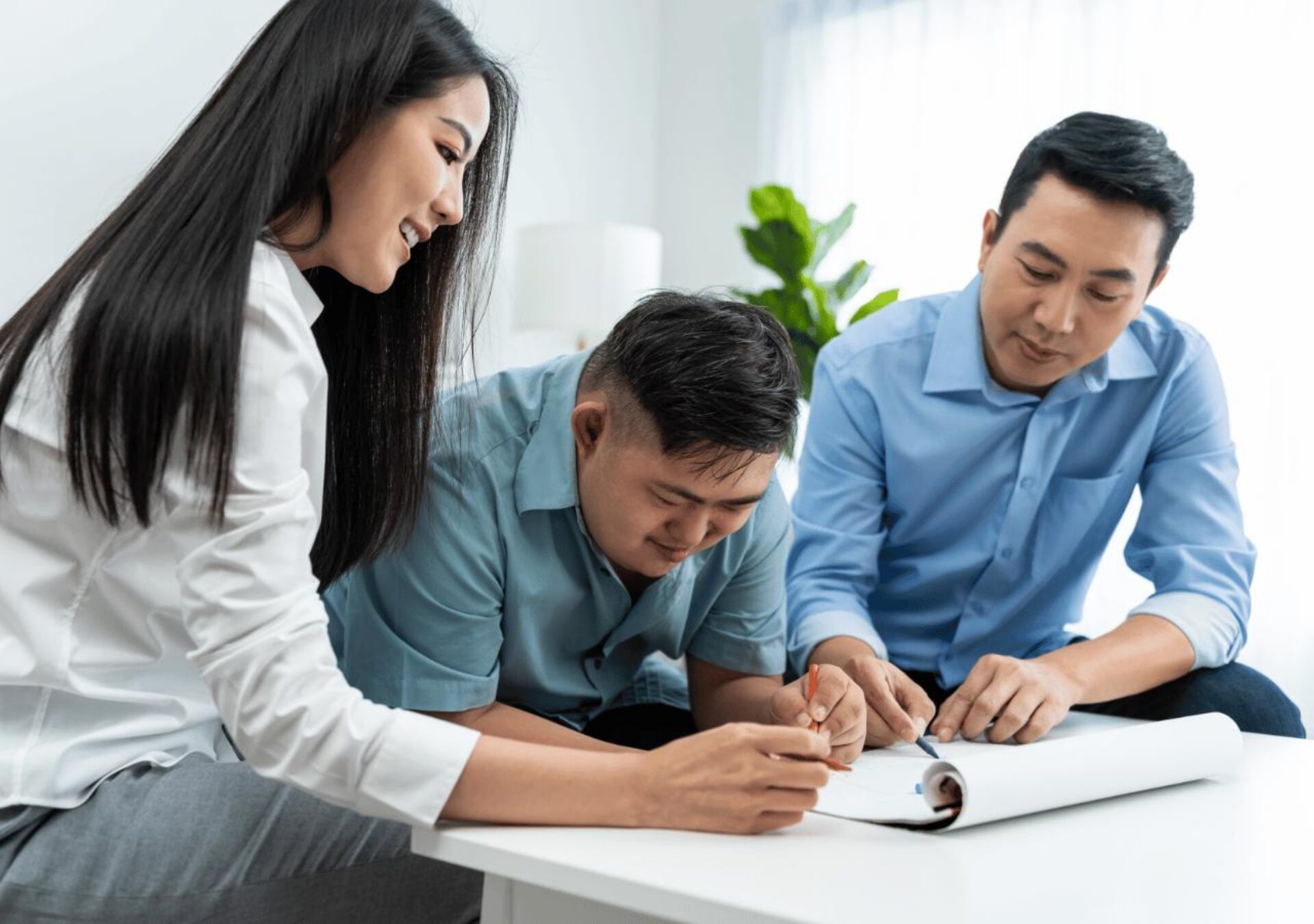 Three business professionals collaborate around a white table, with a woman in a white blazer and two men in blue shirts reviewing documents together in a bright, modern office setting.