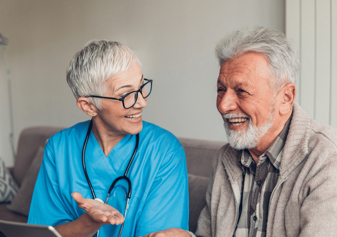 A smiling healthcare worker in blue scrubs and glasses sits beside an elderly man with a grey beard, both laughing together during what appears to be a friendly medical consultation or home visit.