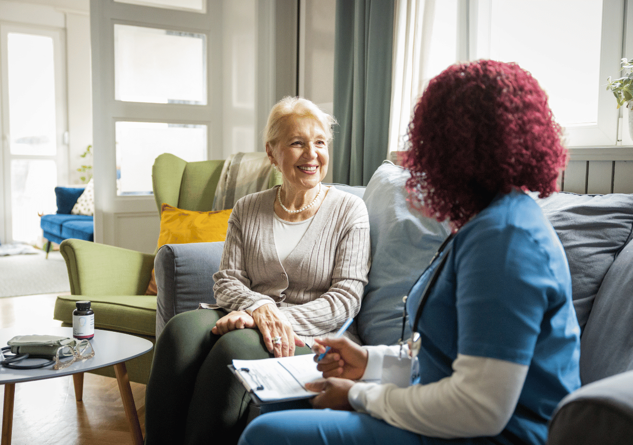 An elderly woman with white hair and pearl necklace sits smiling on a gray couch while speaking with a healthcare worker in blue scrubs who is holding a clipboard, in a bright living room setting.