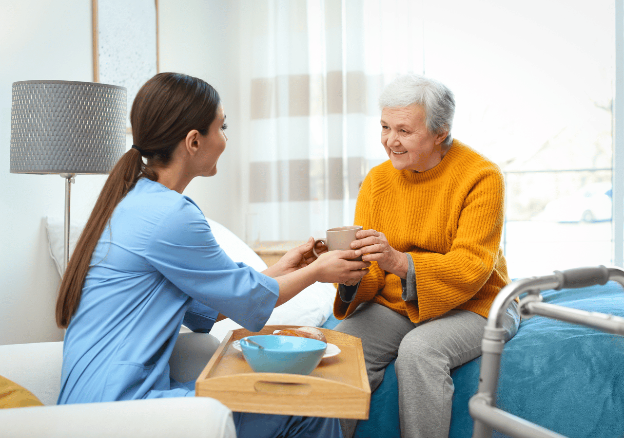A healthcare worker in blue scrubs assists an elderly woman in an orange sweater with drinking from a cup, sitting together in a bright, modern care facility room with a hospital bed visible nearby.