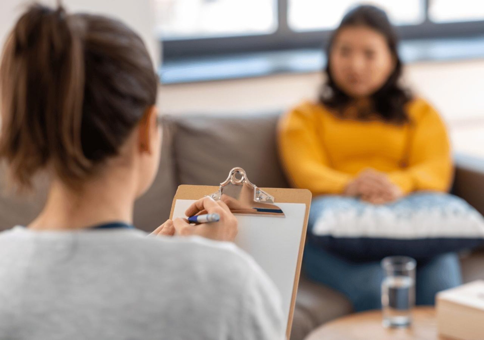 A therapist or counselor takes notes on a clipboard while sitting across from a client in a yellow sweater during a therapy session in a bright, modern office setting.