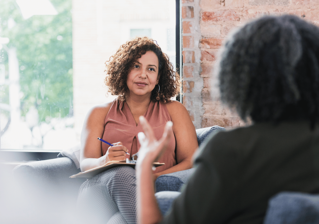 A therapist with curly hair sits attentively on a gray couch taking notes while listening to a client during a therapy session in a modern office with exposed brick walls.