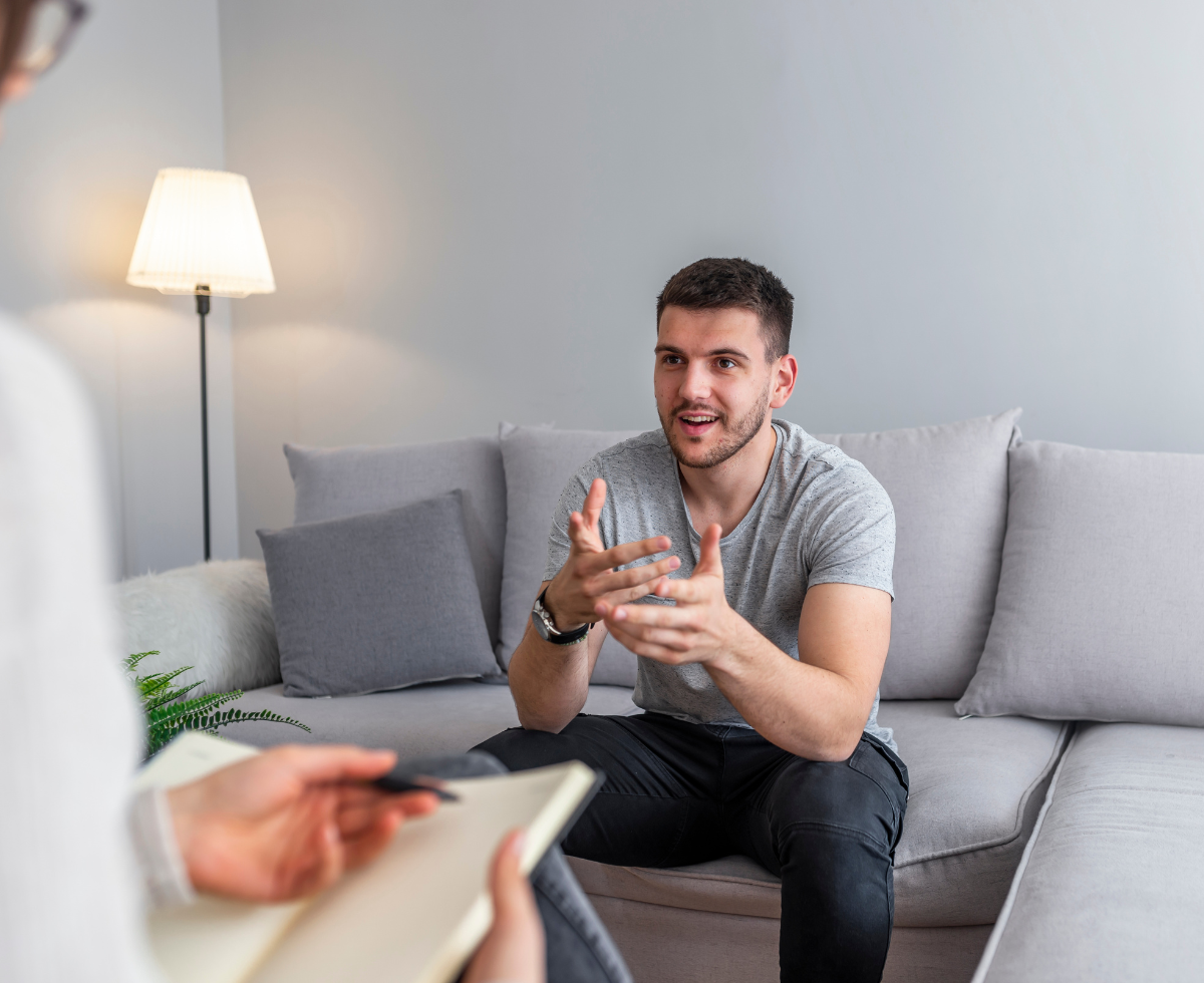 A man in a gray t-shirt sits on a light gray couch gesturing with his hands while speaking to someone across from him who is holding a notebook, suggesting a counseling or interview session in a modern living room setting.