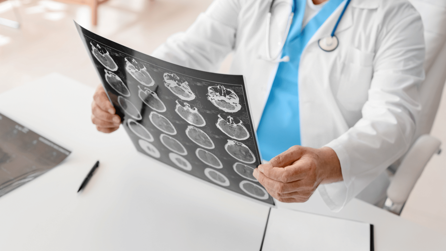 A medical professional in a white coat and blue scrubs examines CT scan images showing cross-sectional views of the brain or head in a clinical setting.