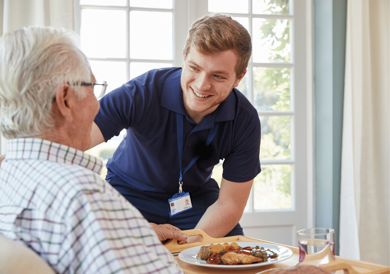 A young male caregiver in a blue polo shirt and ID badge smiles while serving a meal to an elderly man with white hair and glasses at a wooden table near a bright window.