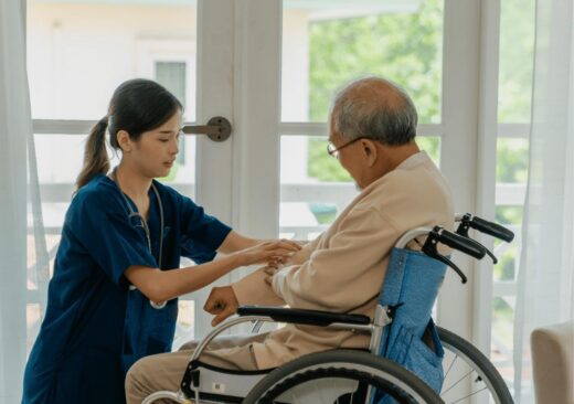 A healthcare worker in blue scrubs gently holds the hands of an elderly man in a wheelchair near a bright window, providing compassionate care in what appears to be a medical or care facility.