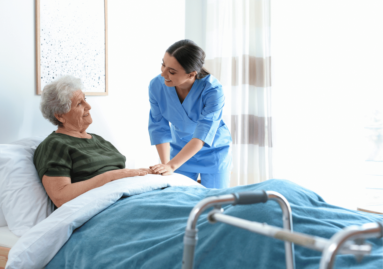 A healthcare worker in blue scrubs provides compassionate care to an elderly patient lying in a hospital bed, gently placing her hands on the patient's arm while they share a warm interaction in a bright medical room.