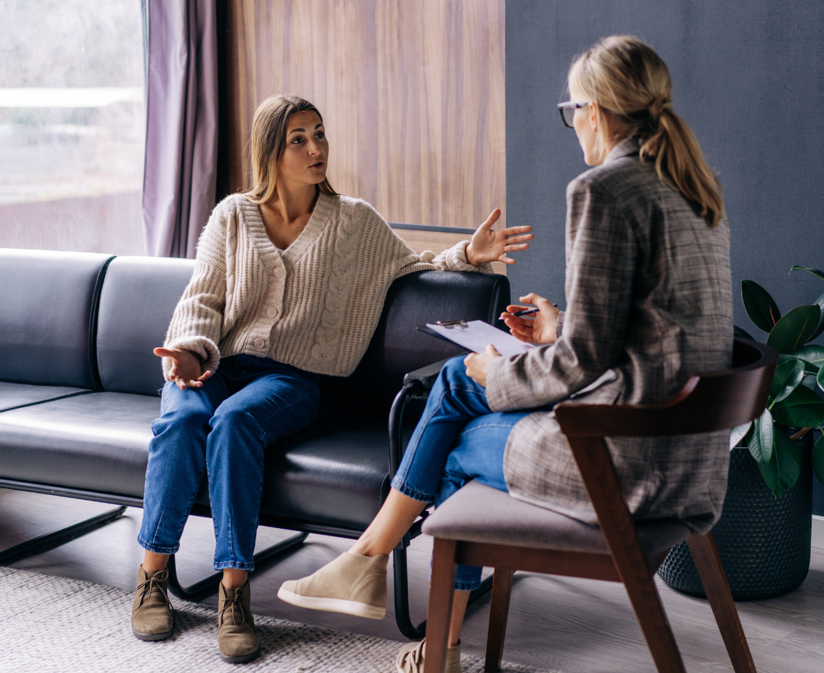 A woman in a cream sweater sits on a gray couch speaking with gesturing hands to a therapist or counselor who sits across from her in a chair, holding a notepad during what appears to be a therapy session in a modern office setting.