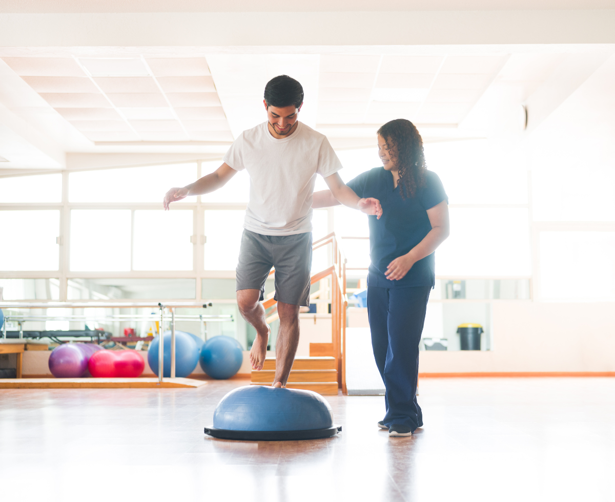 A physical therapist assists a male patient with balance training as he stands on one leg on a blue balance trainer, while she provides support by holding his hand in a bright rehabilitation facility.