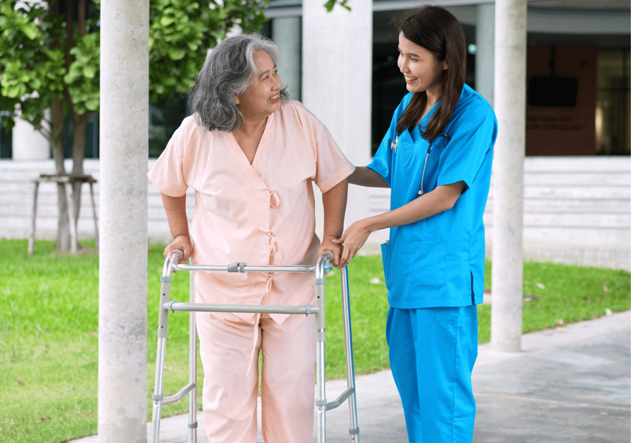 A healthcare worker in blue scrubs assists an elderly woman in pink pajamas who is using a walker, as they walk together on a paved area outside a medical facility or care home.