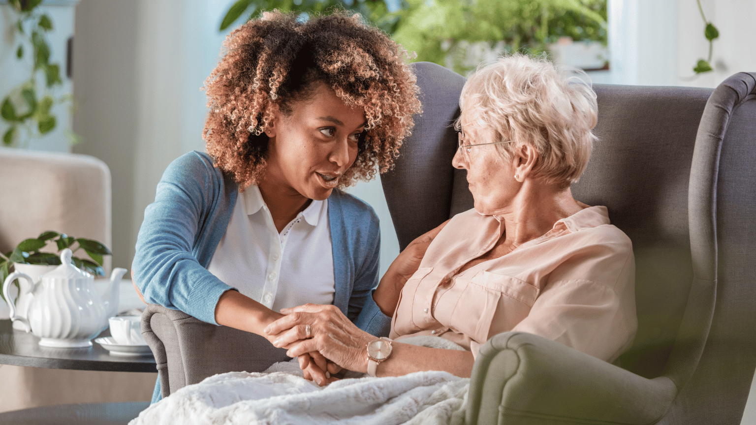 A young healthcare worker with curly hair wearing a blue cardigan sits beside and holds hands with an elderly woman in a peach-colored shirt, both sharing a warm conversation in a comfortable living room setting with plants and tea service visible.