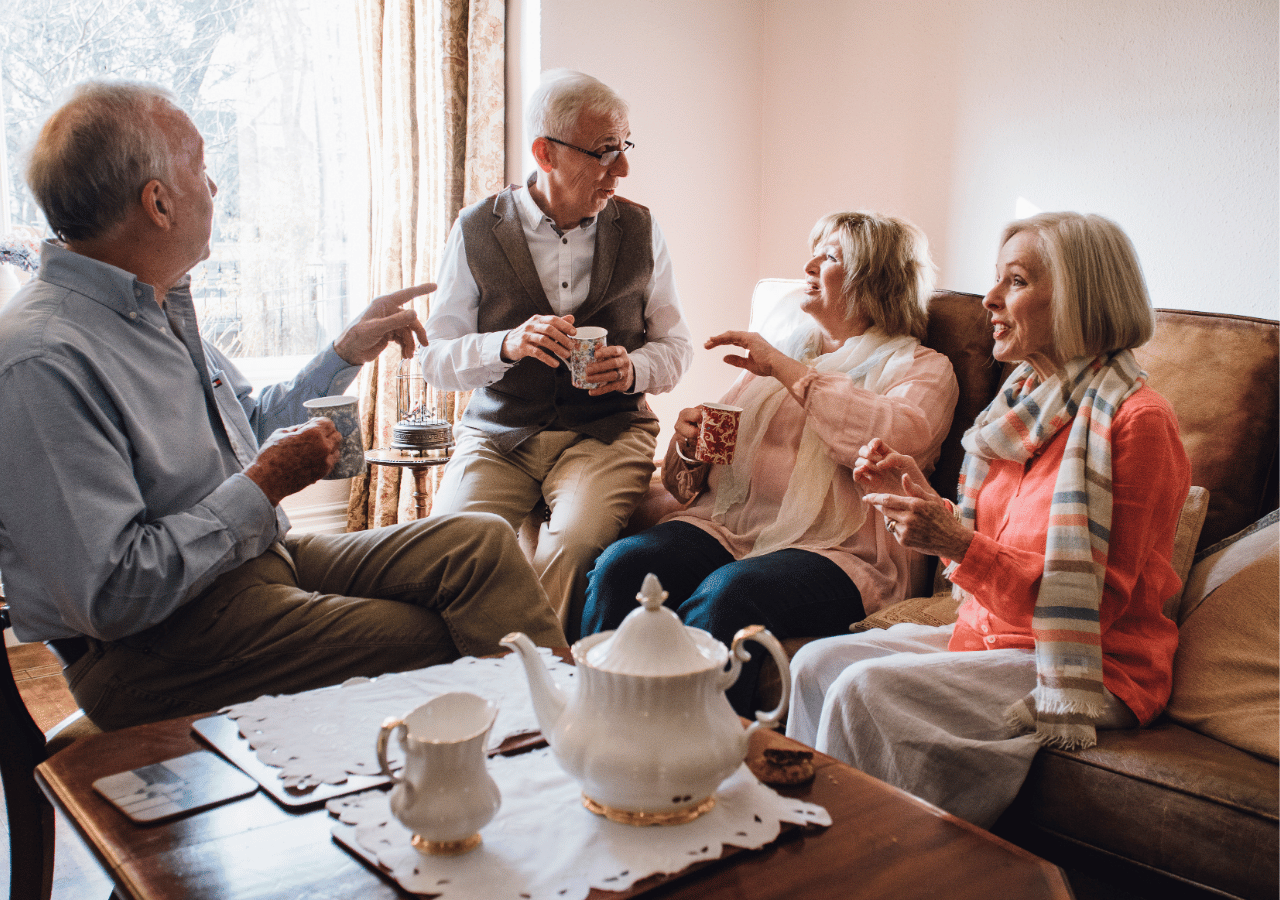 Four older adults sit in a cozy living room having tea and conversation, with a white teapot and cups on the wooden coffee table. The group appears to be enjoying a pleasant social gathering, with warm natural light streaming through the window.