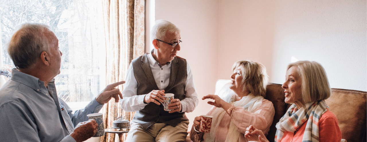 Four older adults enjoy a friendly conversation in a cozy living room, with two men and two women sitting comfortably while holding drinks and engaging in animated discussion near a bright window.