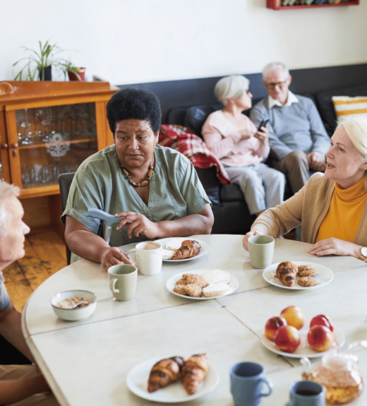 Three elderly people sit around a white table having coffee and pastries in a cozy room with a wooden cabinet and plants, while two other seniors relax on a dark couch in the background.