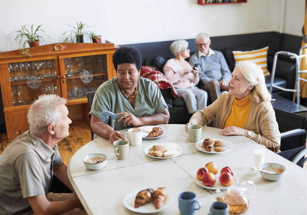 Three elderly people sit around a white table having coffee and pastries in a cozy room with a wooden cabinet and plants, while two other seniors relax on a dark couch in the background.
