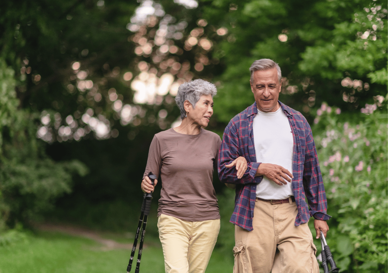 An elderly couple walks together on a path through a lush green park, both using walking sticks for support. The woman wears a brown top and beige pants while the man is dressed in a plaid shirt over a white t-shirt with khaki pants.