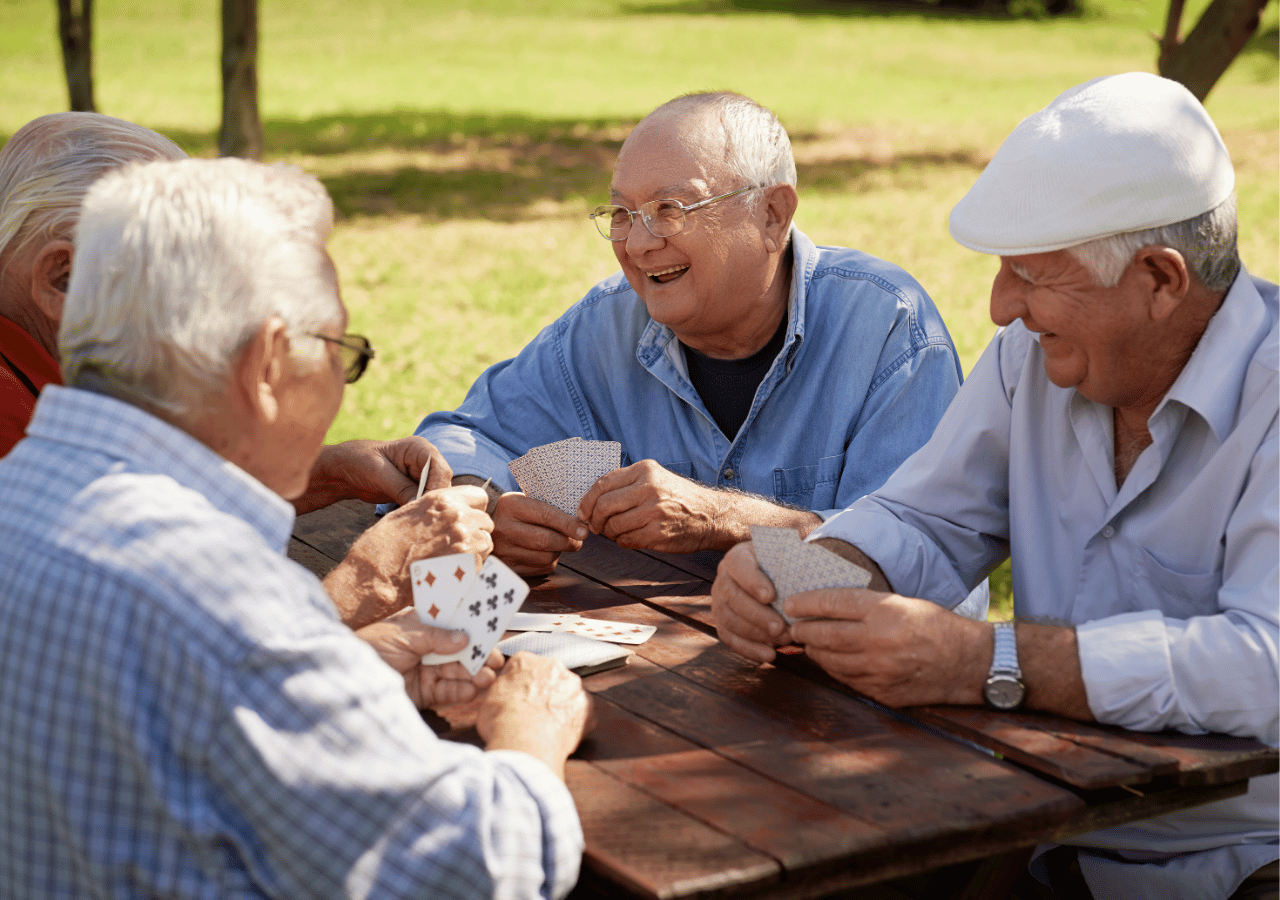 Four elderly men sit around a wooden picnic table playing cards outdoors in a park setting, with one man wearing a white cap and another in glasses and a blue shirt smiling as they enjoy their game together.