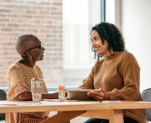 Two women sit at a wooden table in conversation, with one wearing a patterned top and glasses while the other wears a brown sweater and holds a tablet, suggesting a casual meeting or consultation in a bright, modern space.