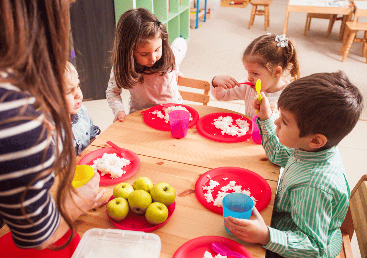 Children sit around a wooden table engaged in a hands-on learning activity, using colorful plates, cups, and what appears to be a white substance like rice or snow, with green apples visible on the table. An adult supervisor stands nearby observing the educational activity in what looks like a classroom or daycare setting.