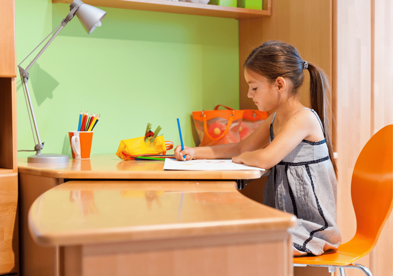 A young girl with a ponytail sits at a wooden desk writing or drawing with a blue pen on white paper. The colorful workspace features a desk lamp, pencil holders, and school supplies, with bright green and orange walls creating a cheerful study environment.