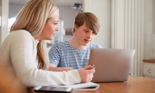 A woman in a white sweater sits beside a teenager in a striped shirt as they look at a laptop together at a wooden table, appearing to be engaged in learning or homework assistance.
