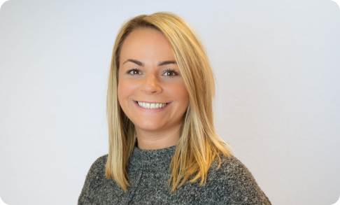 A smiling woman with blonde hair wearing a gray textured sweater poses against a light gray background in what appears to be a professional headshot.
