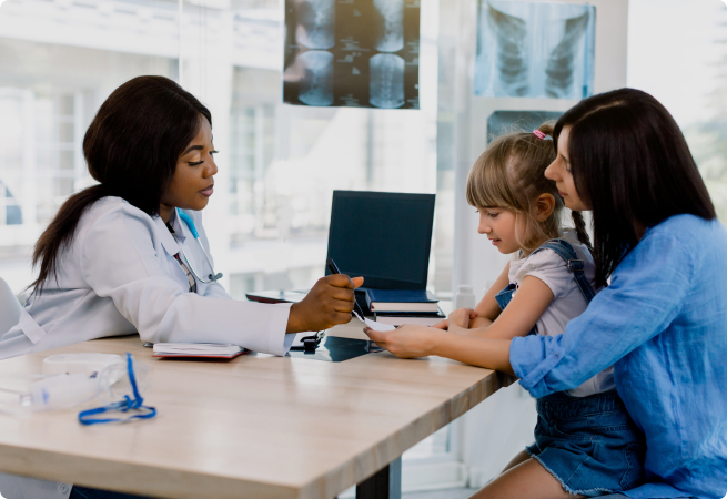 A doctor in a white coat consults with a mother and young child in a medical office, with X-ray images visible on the wall behind them.