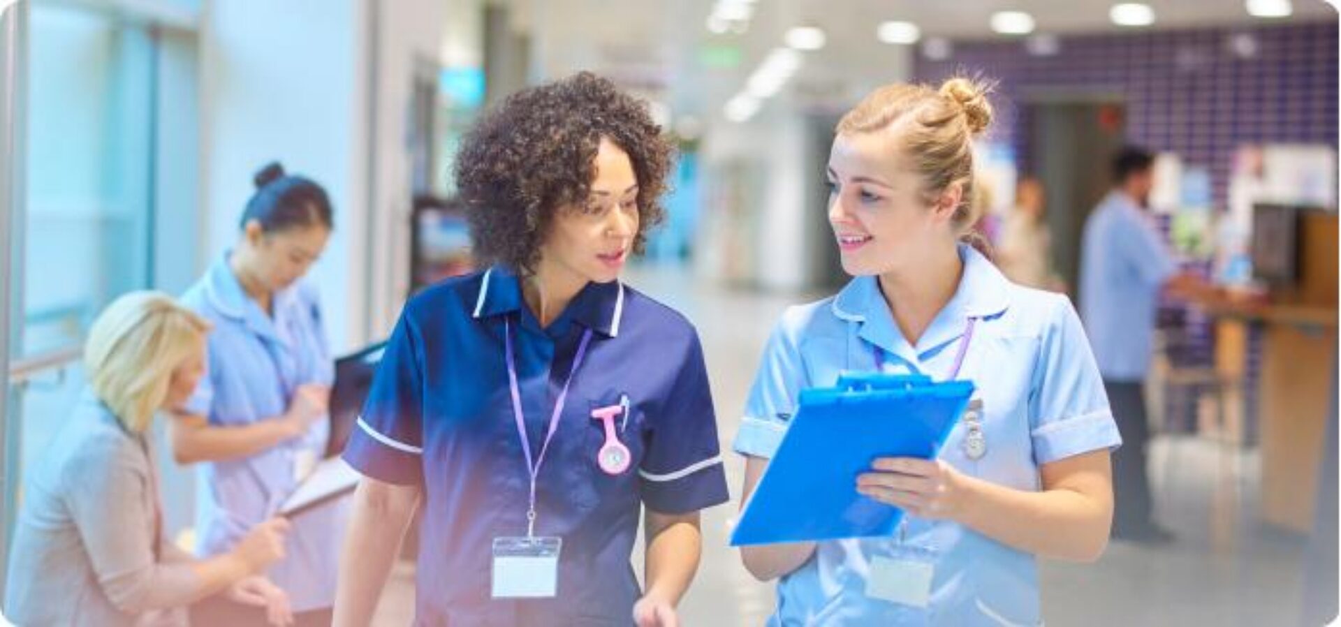 Two healthcare professionals in scrubs walk through a hospital corridor having a conversation, with one holding a blue clipboard, while other medical staff work in the background.