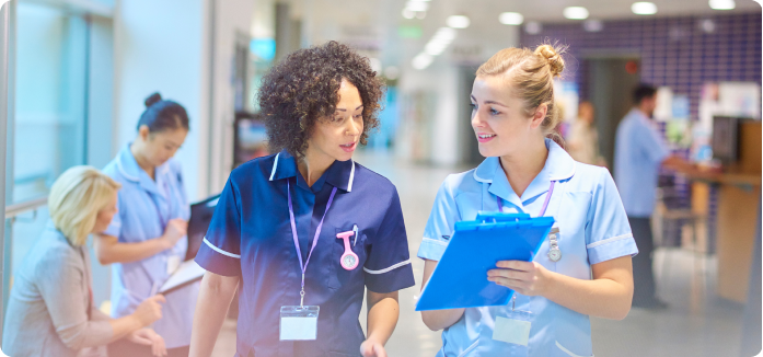 Two healthcare professionals in scrubs walk through a hospital corridor having a conversation, with one holding a blue clipboard, while other medical staff work in the background.