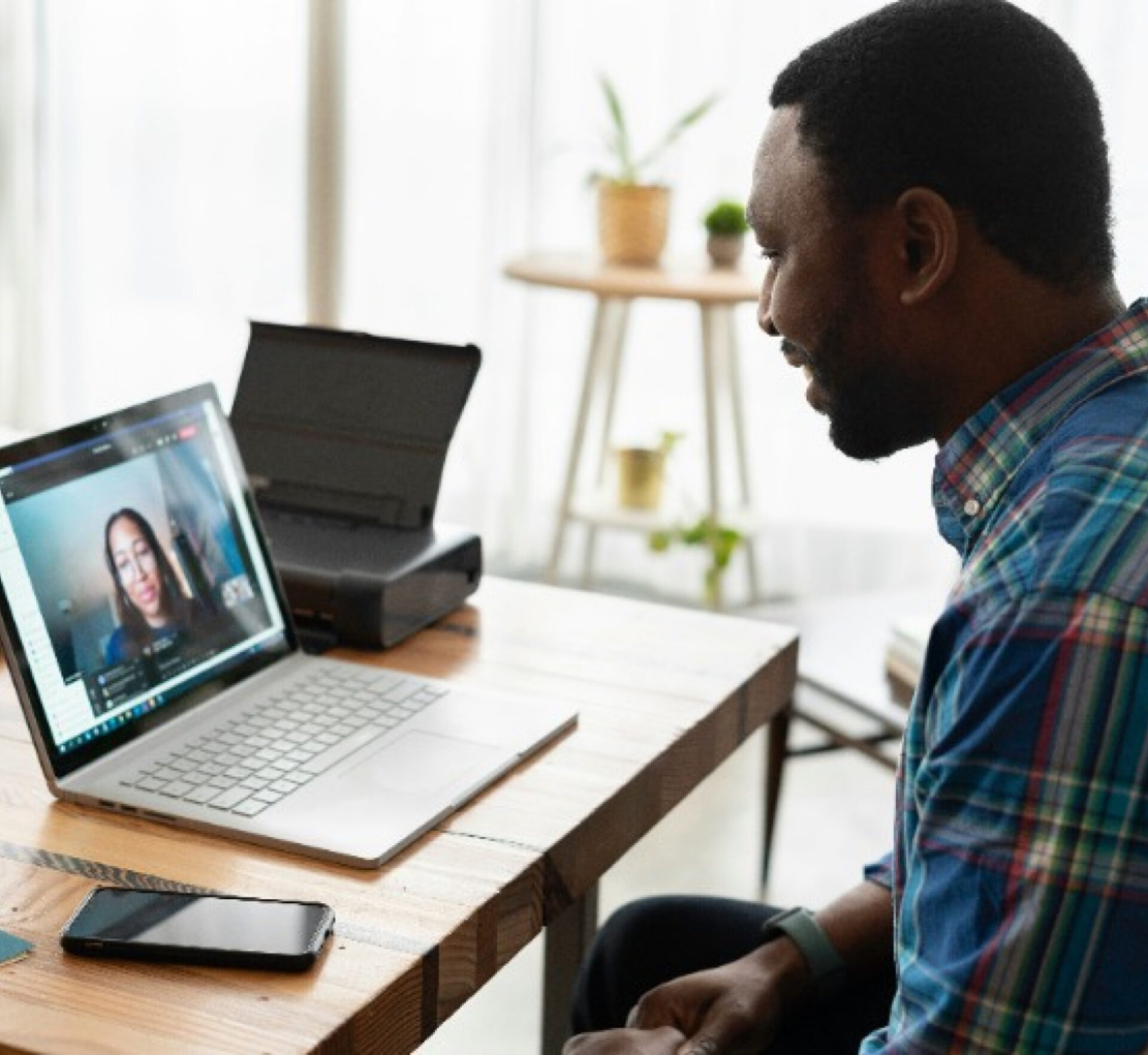 A man in a plaid shirt sits at a wooden desk participating in a video call on his laptop, with a woman visible on the screen and a smartphone placed nearby on the desk.
