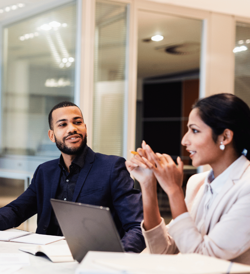 Two business professionals have a discussion at a modern office table, with a man in a dark suit jacket gesturing while speaking and a woman in a white shirt listening attentively, a laptop and documents visible on the table between them.