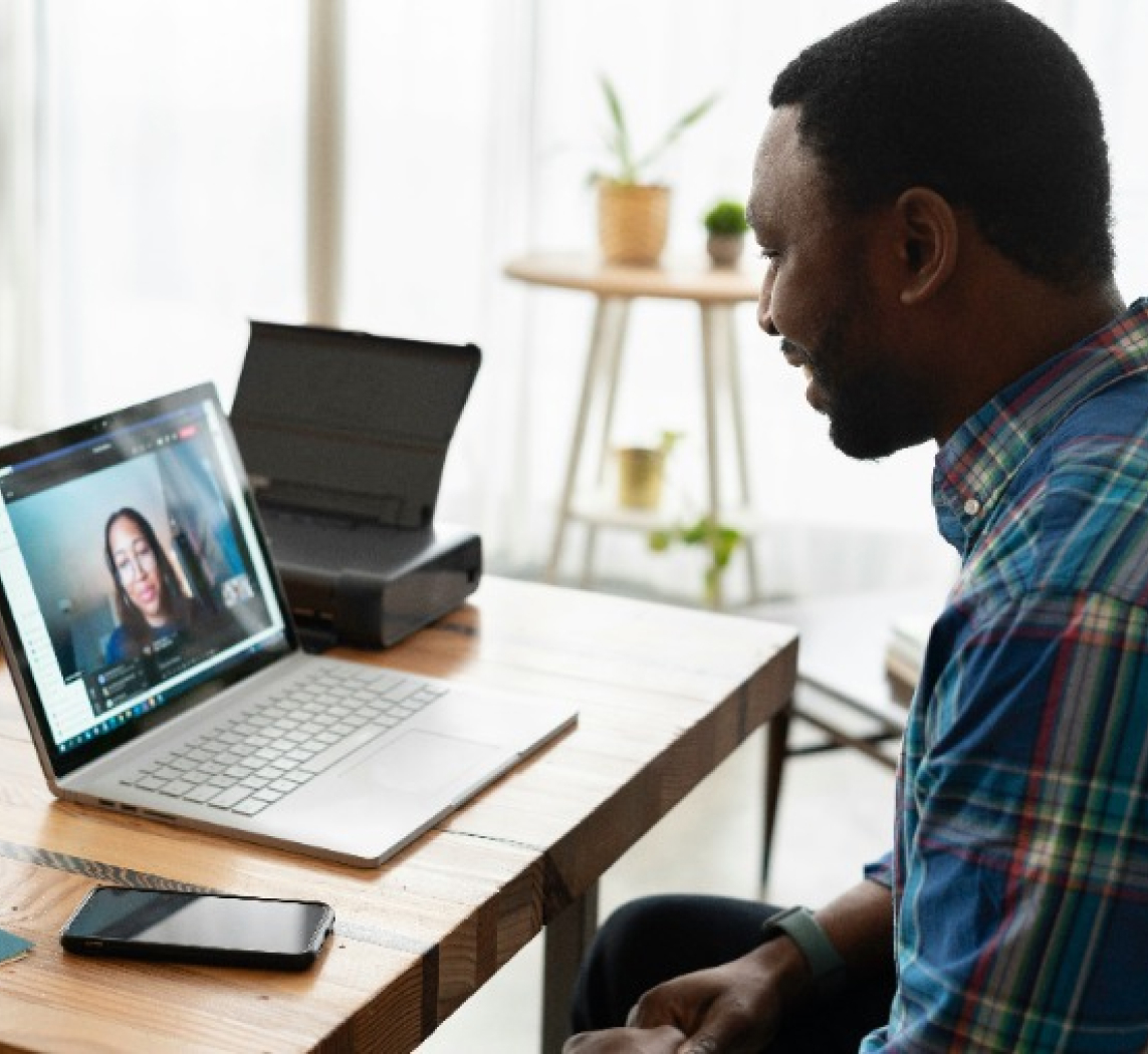 A man in a plaid shirt sits at a wooden desk participating in a video call on his laptop, with a woman visible on the screen and a smartphone placed nearby on the desk.